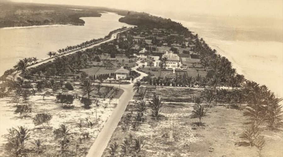 A vintage aerial view of a narrow peninsula lined with palm trees, roads, and scattered buildings, surrounded by water on both sides.