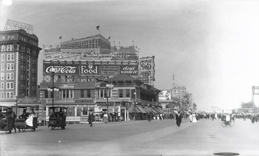 Black and white photo of a busy boardwalk lined with old cars, people in early 1900s attire, and large advertisements for Coca-Cola and other businesses on buildings.