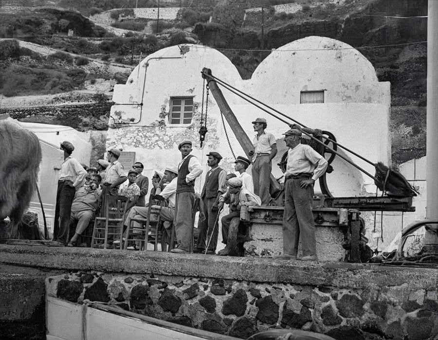 A group of men in work clothes gather near a stone building and a small crane, with a hilly landscape in the background. Most are standing, some sit on chairs, and all appear to be engaged in conversation or observing something.