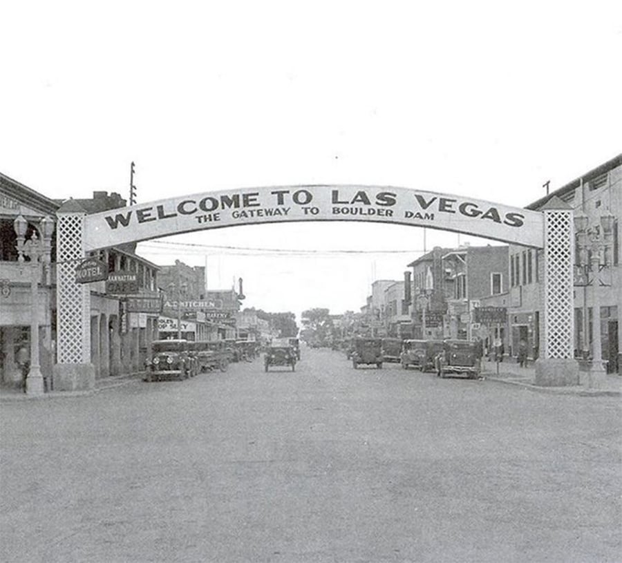 Black-and-white photo of a vintage street scene in Las Vegas, featuring an archway reading “Welcome to Las Vegas, The Gateway to Boulder Dam,” with old cars and shops lining the street.