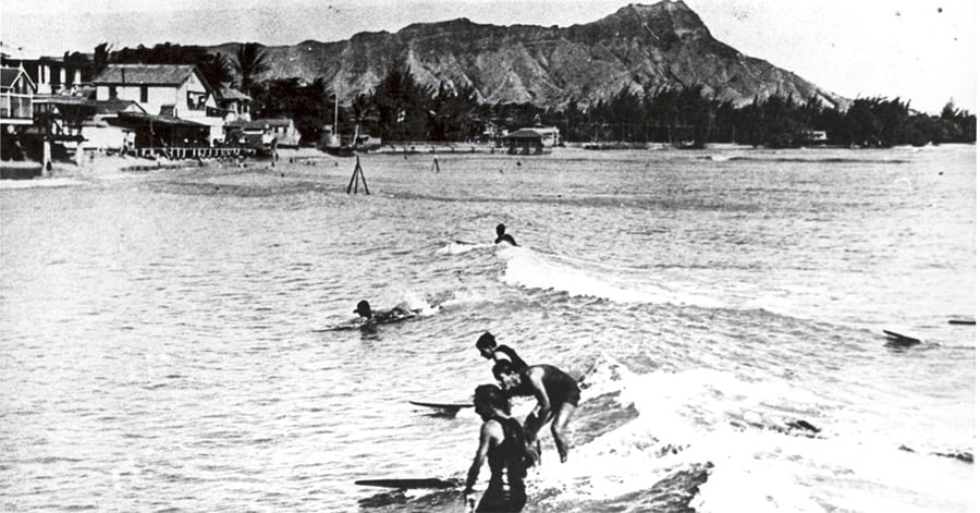Black-and-white photo of several surfers riding small waves near the shore, with houses and a large mountain in the background.