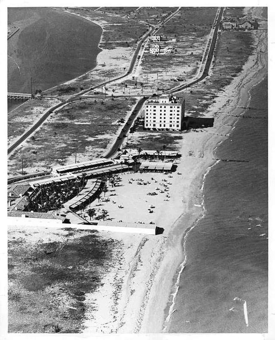 A black-and-white aerial photo of a beachfront hotel with people sunbathing on the sand, a few buildings nearby, sparse roads, and the shoreline stretching along the right side.