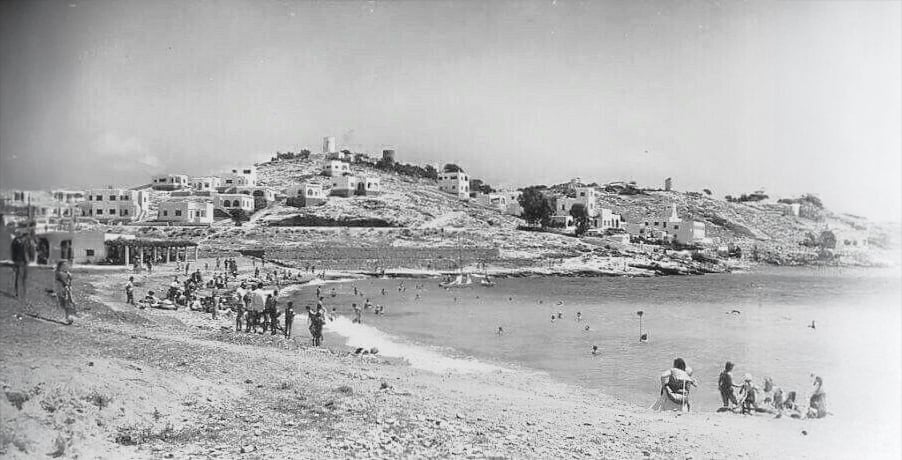 Black-and-white photo of a beach crowded with people swimming and sunbathing. In the background, white buildings and windmills are scattered on a hillside under a clear sky.