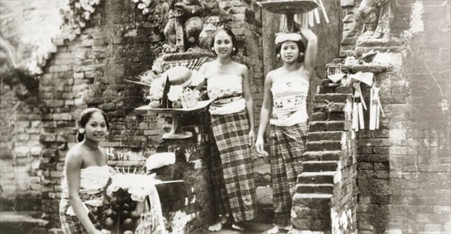 Three women in traditional attire stand by stone ruins, holding and carrying baskets of offerings. One is on a step, another stands beside her, and the third kneels in front. The scene appears historical and ceremonial.