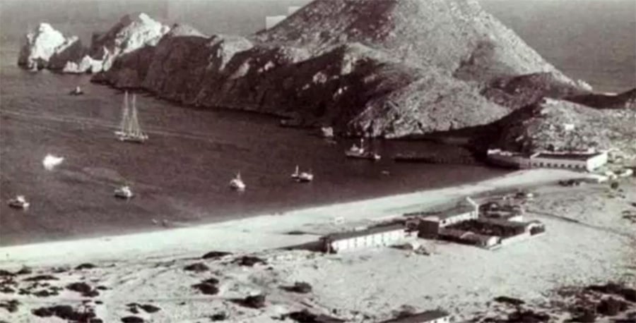 A black-and-white aerial photo shows a rocky coastline with a few boats in the water, a sandy beach, and sparse buildings near the shore, backed by rugged hills.