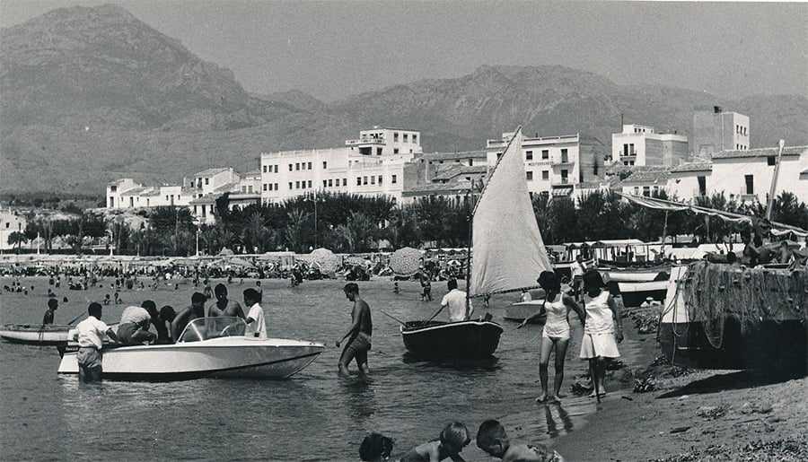 People gather on a beach with boats in the water and mountains in the background. Buildings line the shore, and some people are wading or standing near the boats. The photo appears to be from a past era.
