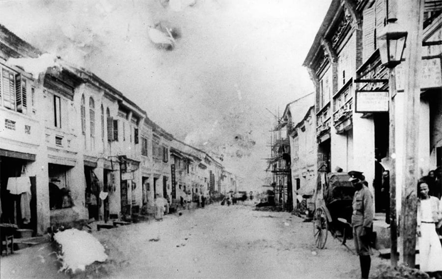 Black-and-white photo of an old street with colonial-style buildings, people walking, and a few standing; some scaffolding is visible on the right. The scene appears from the early 20th century.