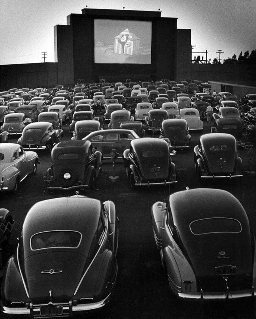 Black and white photo of cars parked at a drive-in theater at night, facing a large outdoor screen showing a movie with a person in robes on it. The cars are from the mid-20th century.