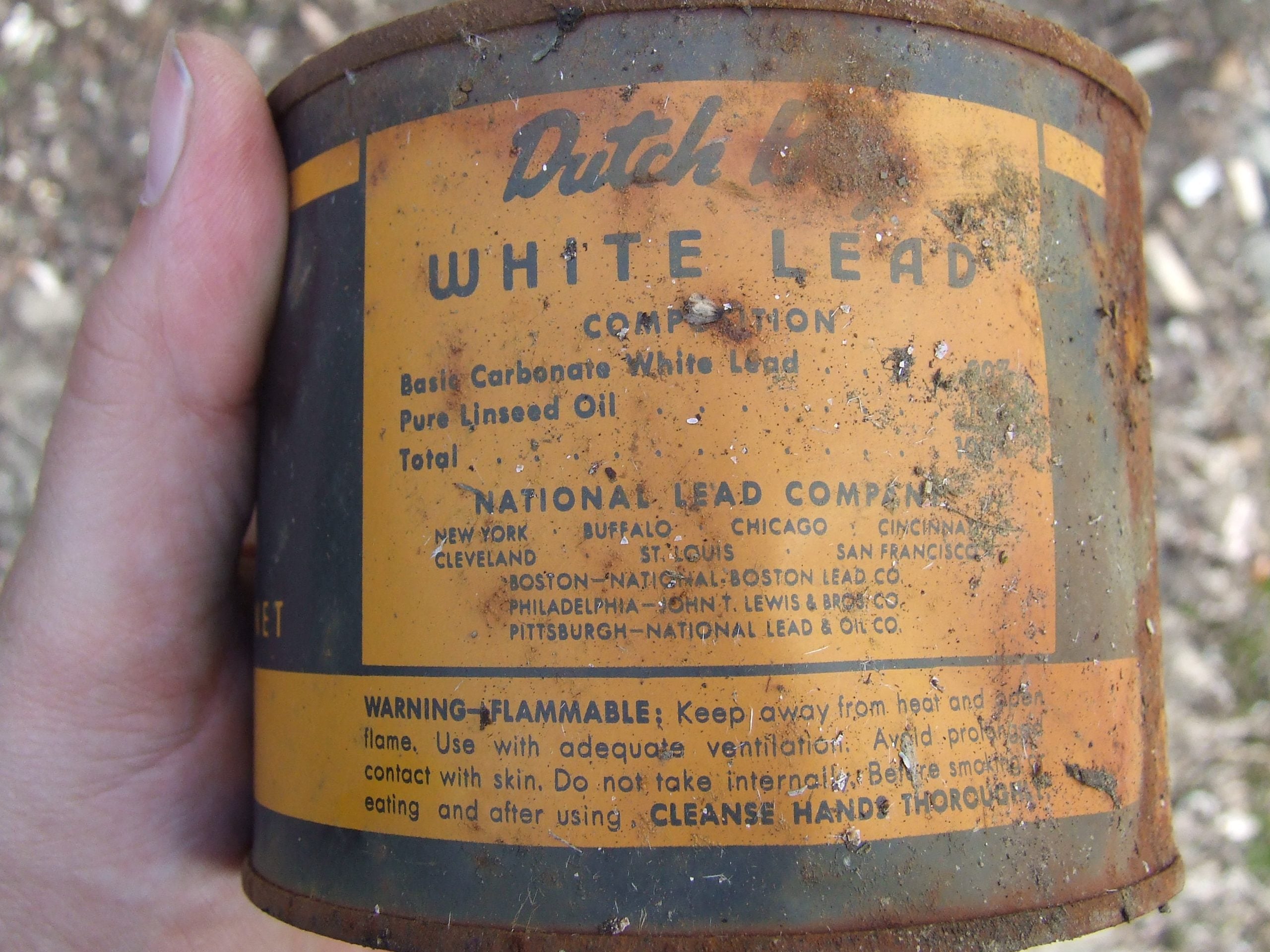A hand holds a rusty can labeled “Dutch Boy White Lead.” The label lists ingredients, warnings about lead exposure, and manufacturers. The can appears old and weathered.