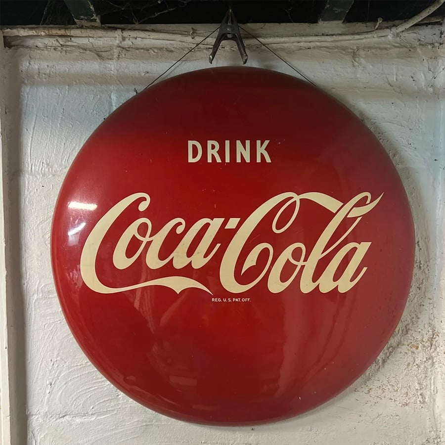 A vintage red Coca-Cola sign with white lettering reading “Drink Coca-Cola,” hanging on a white textured wall.
