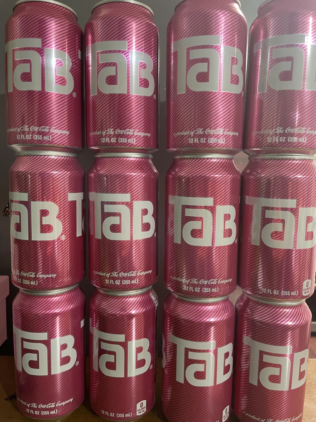 Twelve pink cans of TaB diet soda are stacked in three rows of four. The cans feature the TaB logo in large white letters on a diagonal striped background.
