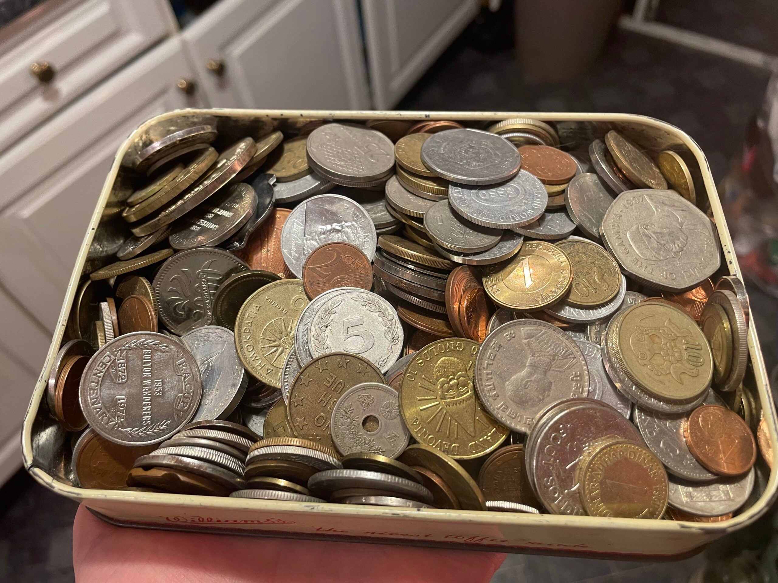 A hand holds an open metal tin filled with assorted coins of various sizes, colors, and countries, with kitchen cabinets and a trash bin visible in the blurry background.