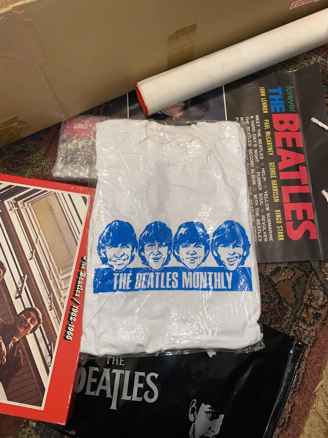 A white T-shirt with The Beatles’ faces in blue is wrapped in plastic, surrounded by Beatles magazines, books, and memorabilia on a patterned surface.