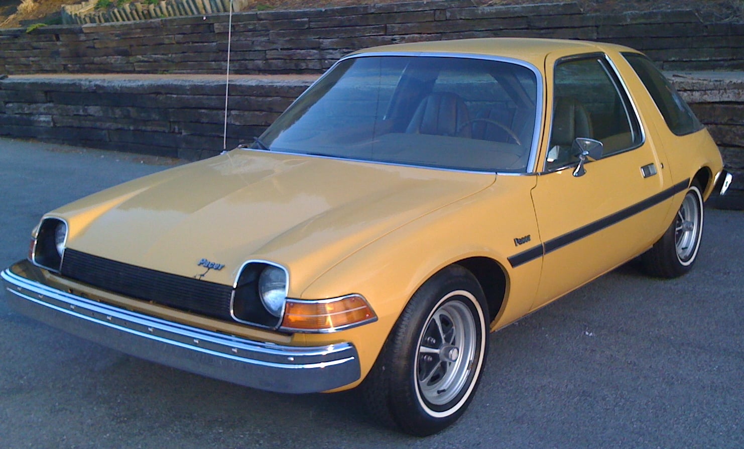 A yellow classic two-door car is parked on a paved surface near a stone wall, with “Pacer” written on the side.
