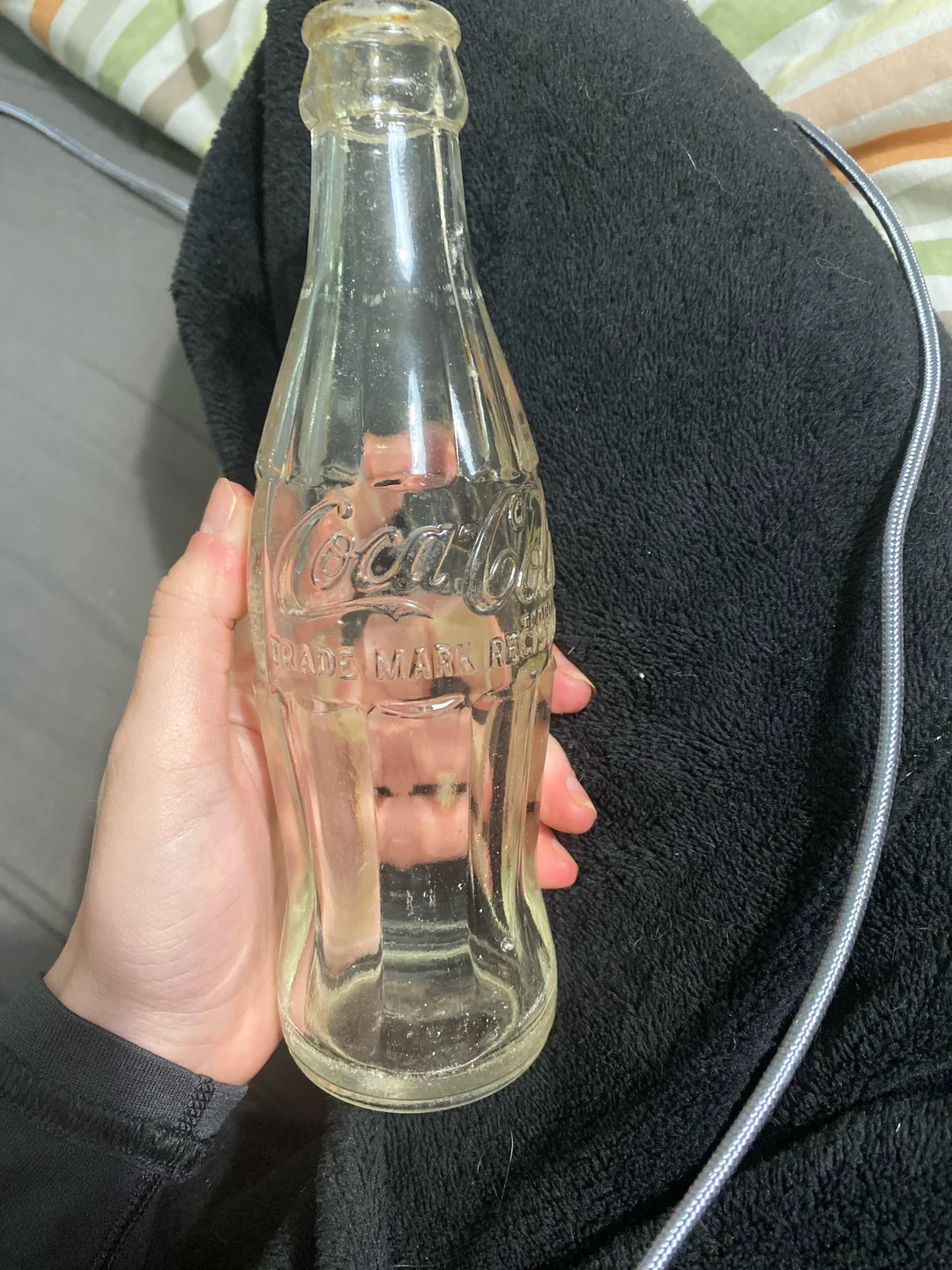 A hand holds a vintage, clear glass Coca-Cola bottle with “Coca-Cola” embossed on it. The bottle is empty and resting on a black fabric surface. A cord and striped bedding are partially visible in the background.