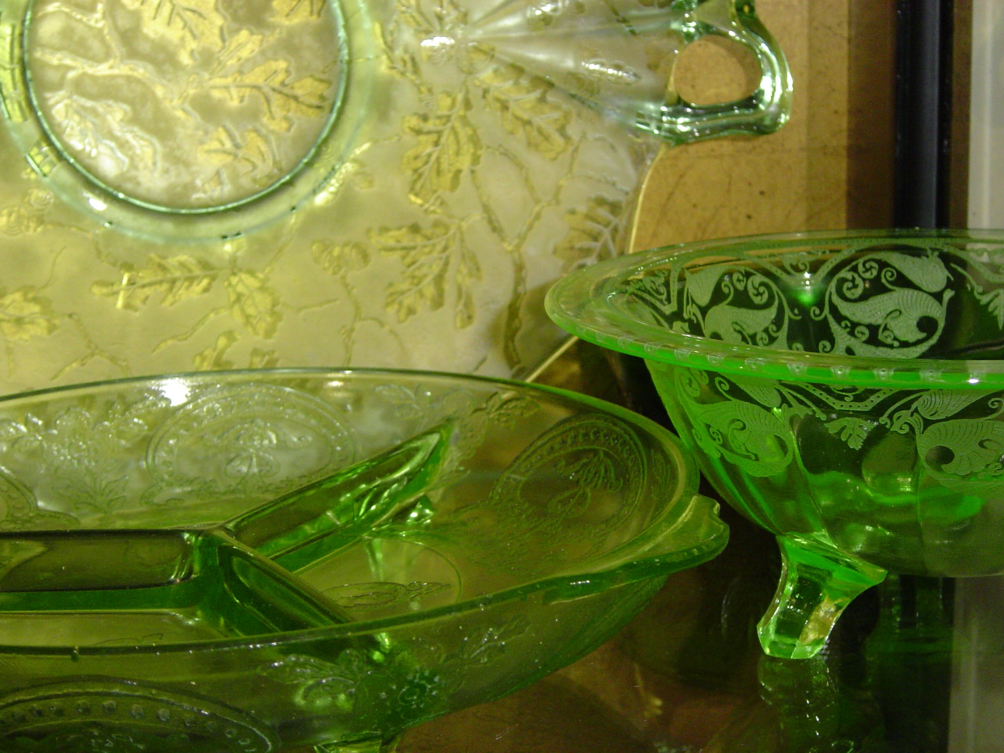 Green Depression glass dishes with ornate patterns are displayed, including a sectioned platter, a bowl with three feet, and a plate in the background, all with floral and leafy embossed designs.