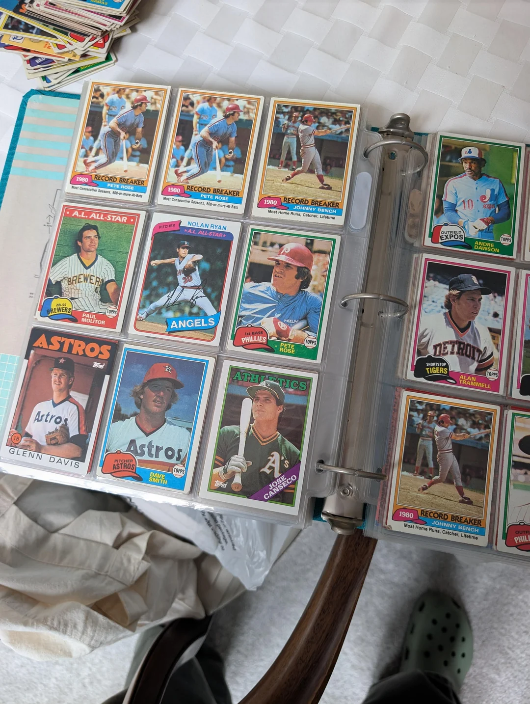 Open binder displaying pages of vintage Topps baseball cards in plastic sleeves, featuring players in various team uniforms. The binder rests on a table, and a person’s feet in green Crocs are visible below.