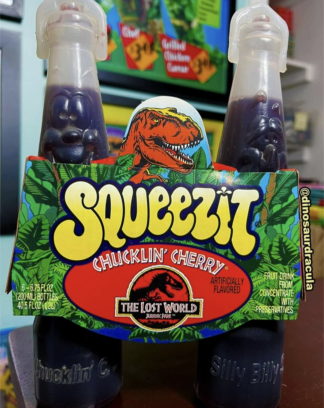 Two purple Squeezit bottles in Chucklin’ Cherry flavor with The Lost World: Jurassic Park branding, featuring a dinosaur illustration on the packaging and a TV showing a related image in the background.