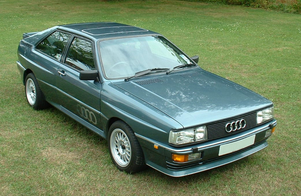 A classic dark green Audi Quattro coupe parked on grass, viewed from above at an angle, showing its boxy shape, alloy wheels, and distinctive grille with four rings.