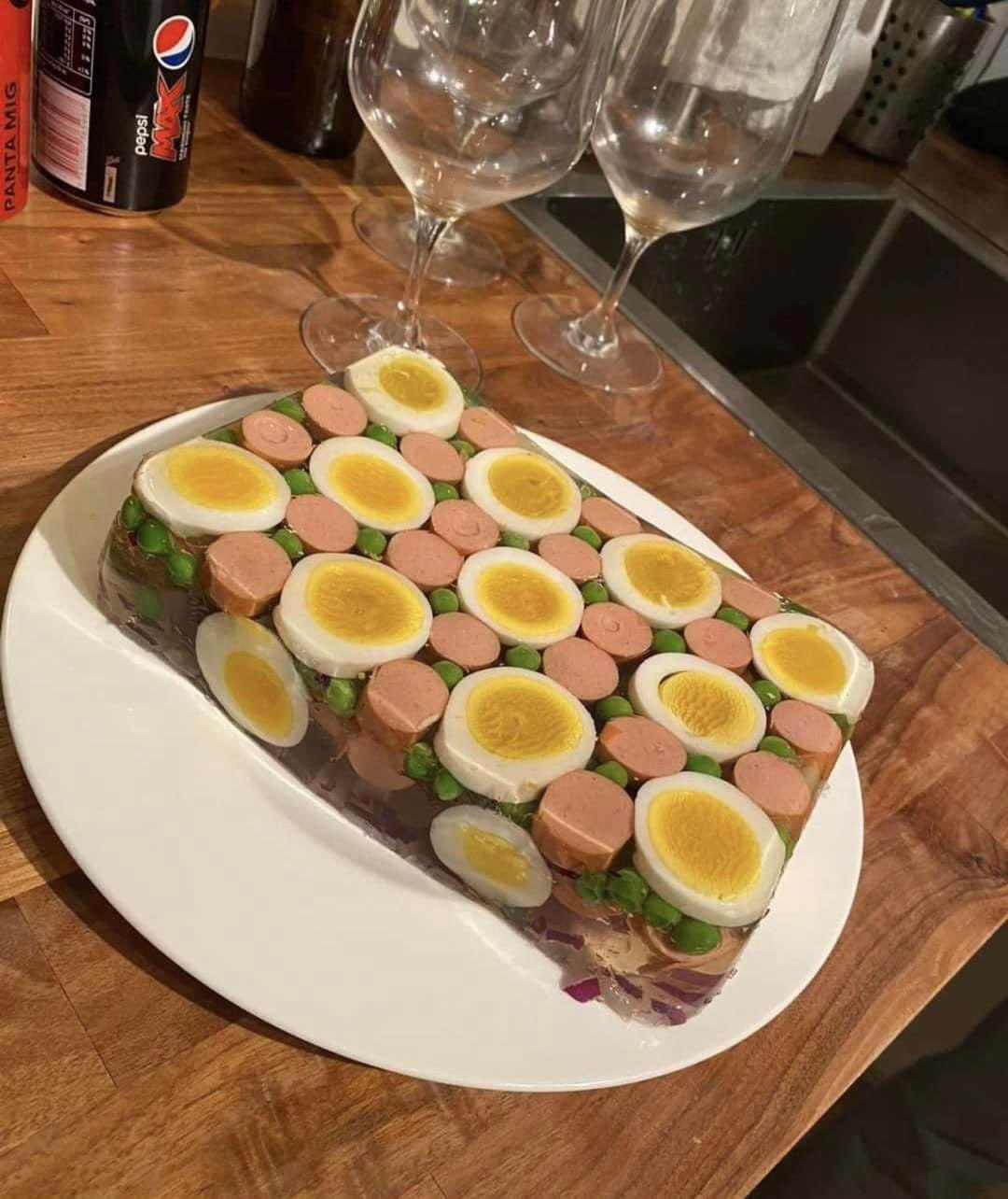 A rectangular gelatin dish with sliced boiled eggs, sausage pieces, and green vegetables, served on a white plate on a wooden counter with glasses and a Pepsi can in the background.