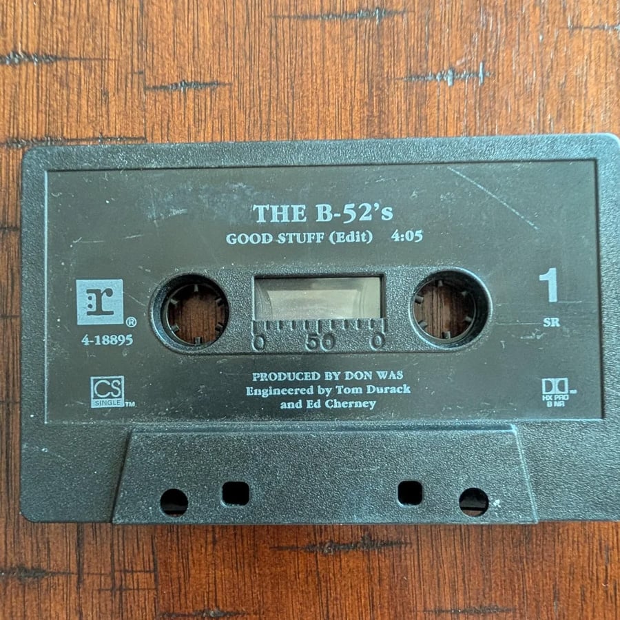 A black cassette tape labeled “THE B-52’s GOOD STUFF (Edit) 4:05” sits on a wooden surface. Production credits and logos are printed on the front of the tape.