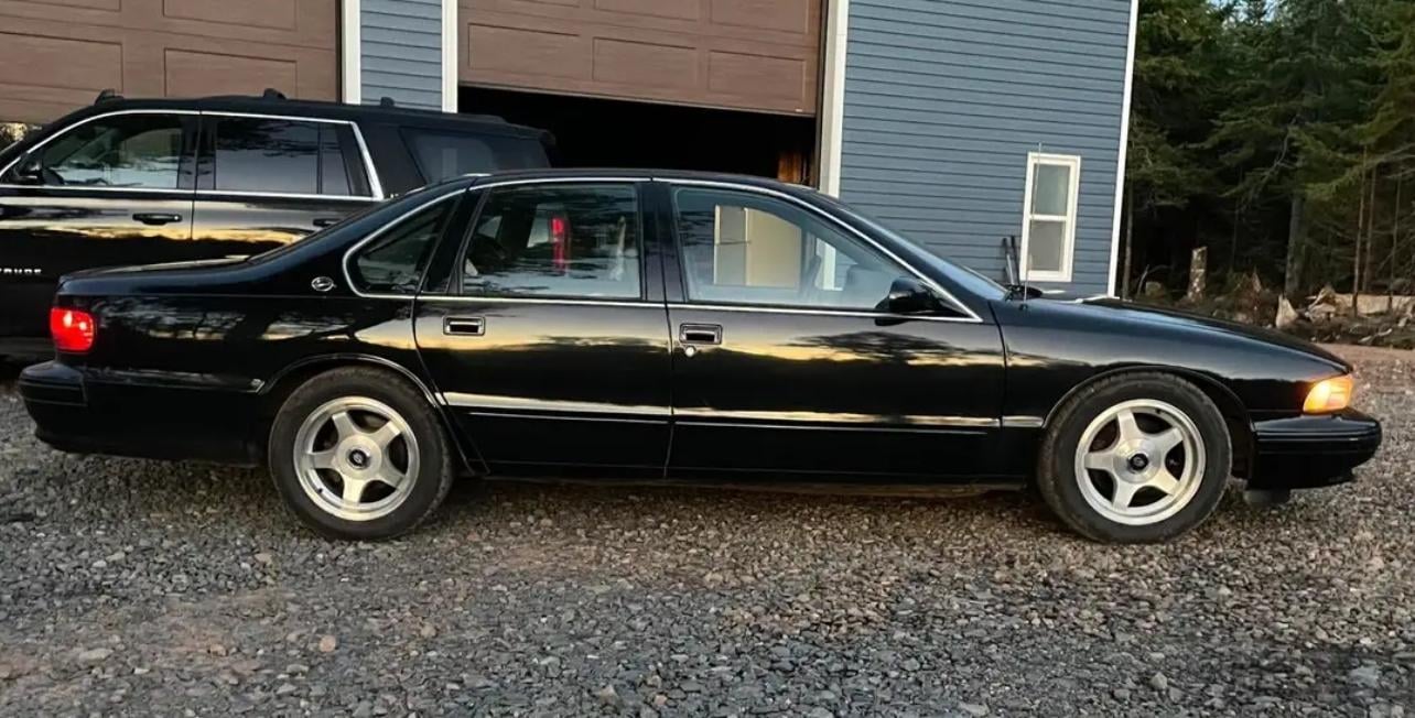 A black sedan with silver rims is parked on gravel in front of a blue building and next to a black SUV. Trees are visible in the background.