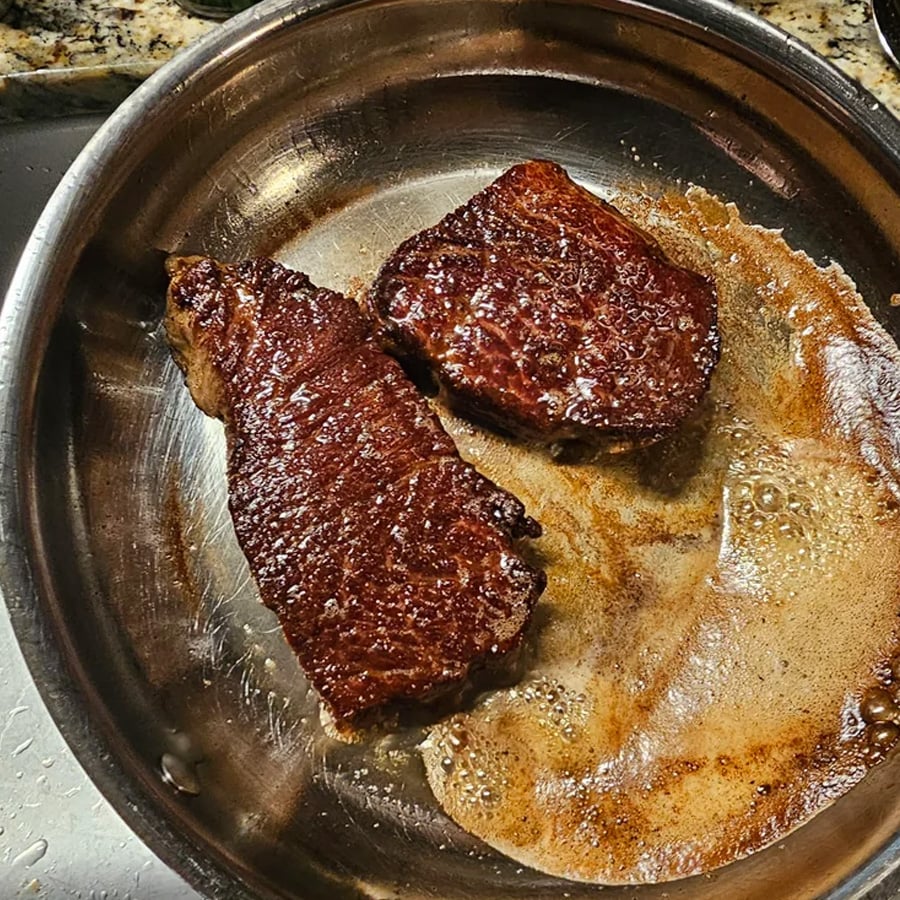 Two seared steaks cooking in a stainless steel pan with bubbling brown butter on the side, sitting on a stovetop.