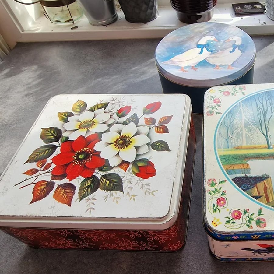 Three decorative metal tins sit on a countertop. One has flowers, another features two geese in bonnets, and the third shows a scenic bridge and cottage illustration. Sunlight streams in from a window behind them.