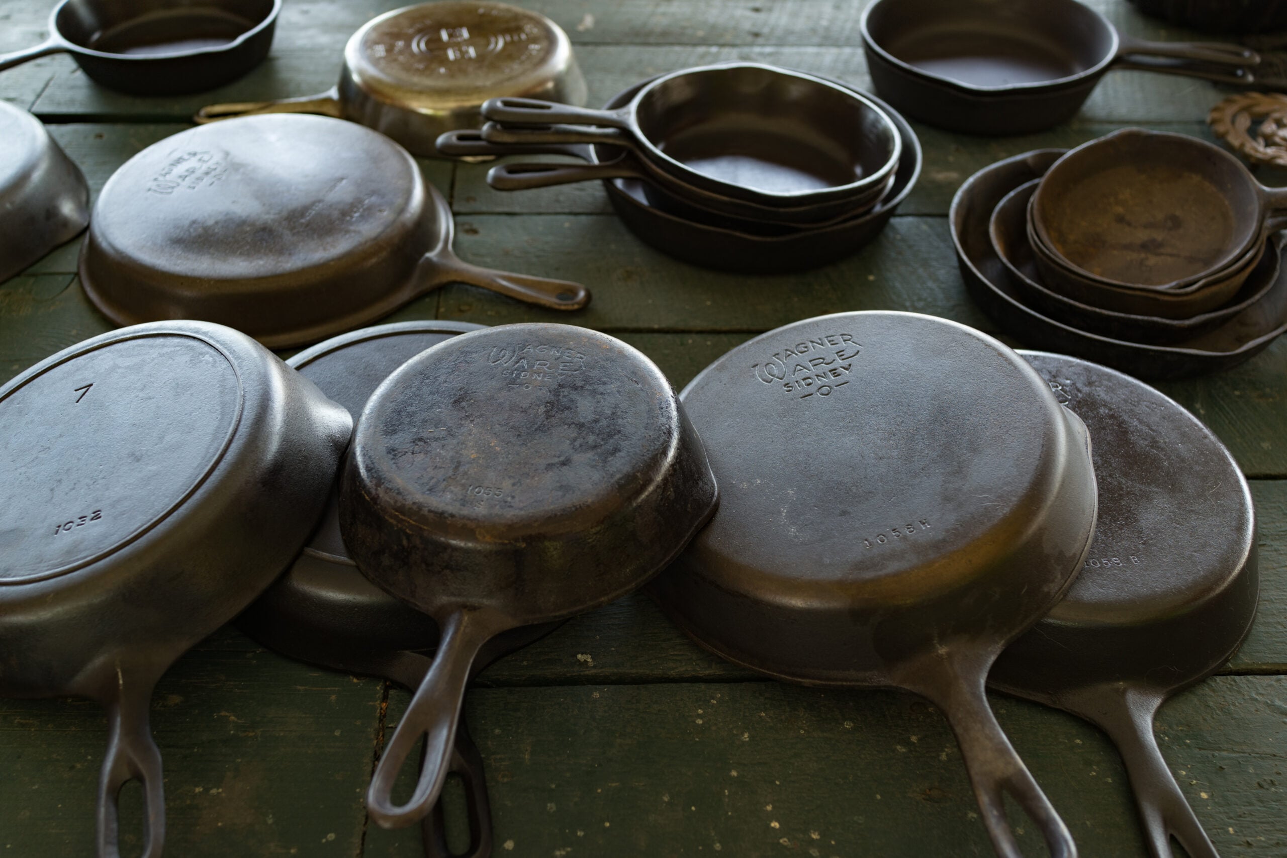 Several cast iron skillets and pans of various sizes are arranged on a rustic green wooden table, some stacked, some placed upside down, showing wear and maker marks.