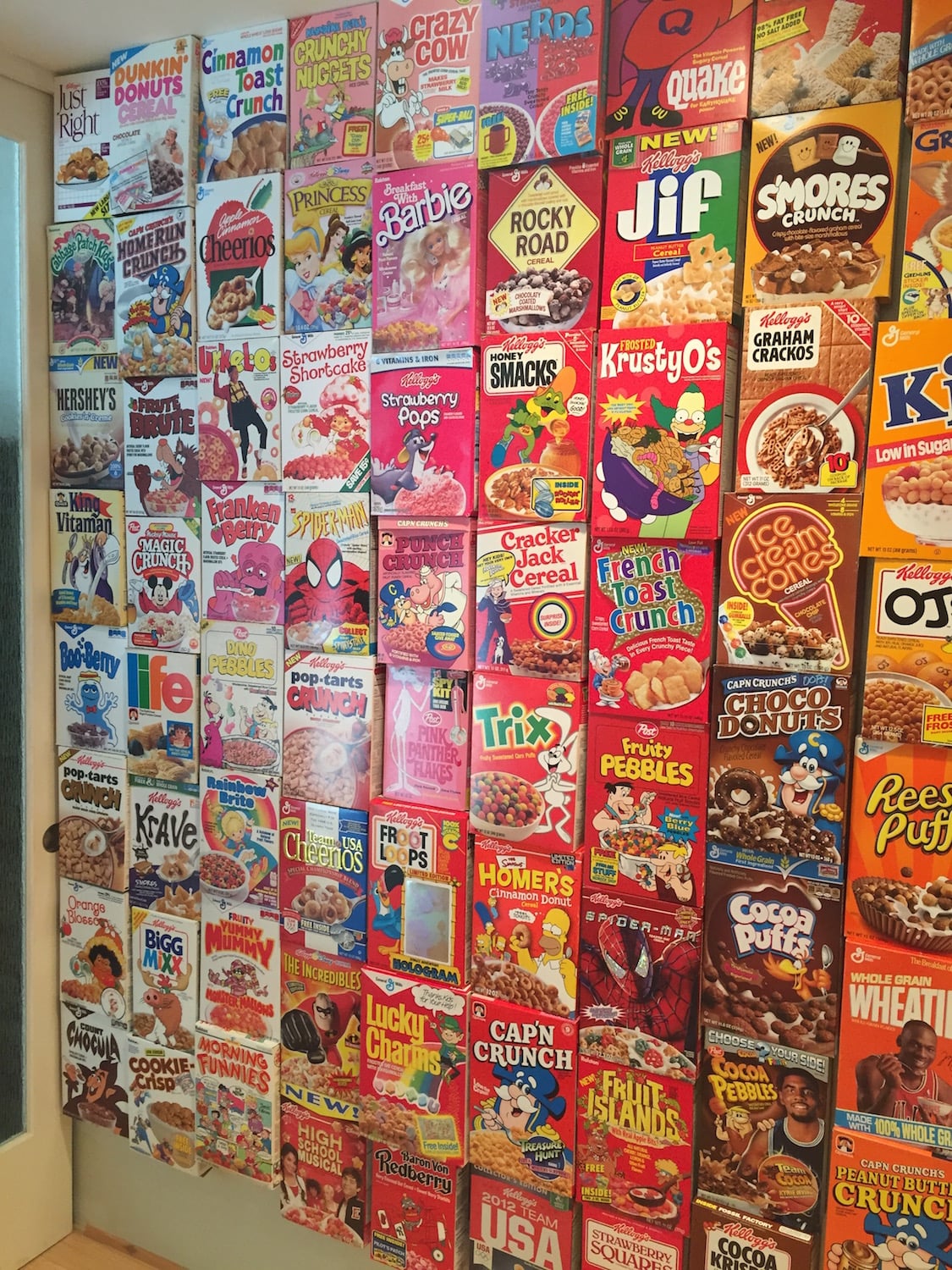 A wall covered with colorful cereal boxes, including brands like Froot Loops, Lucky Charms, Cap’n Crunch, and Cocoa Puffs, arranged closely together in rows.