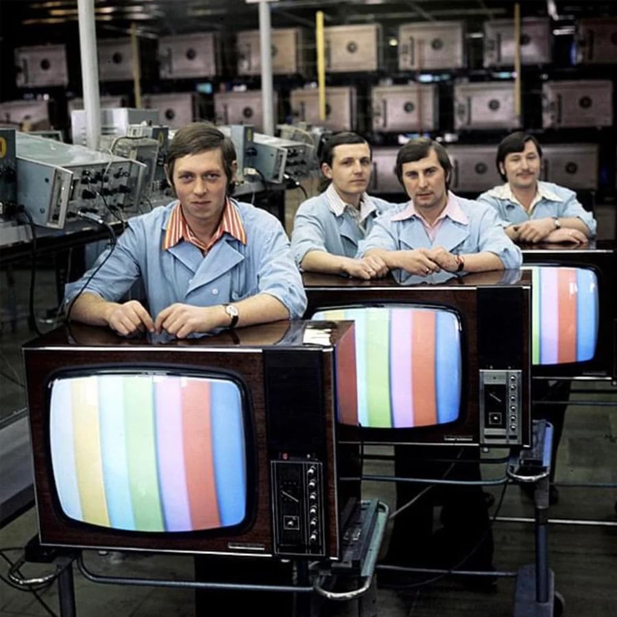 Four men in blue shirts sit behind vintage TVs displaying color test patterns, inside a factory filled with televisions and equipment.