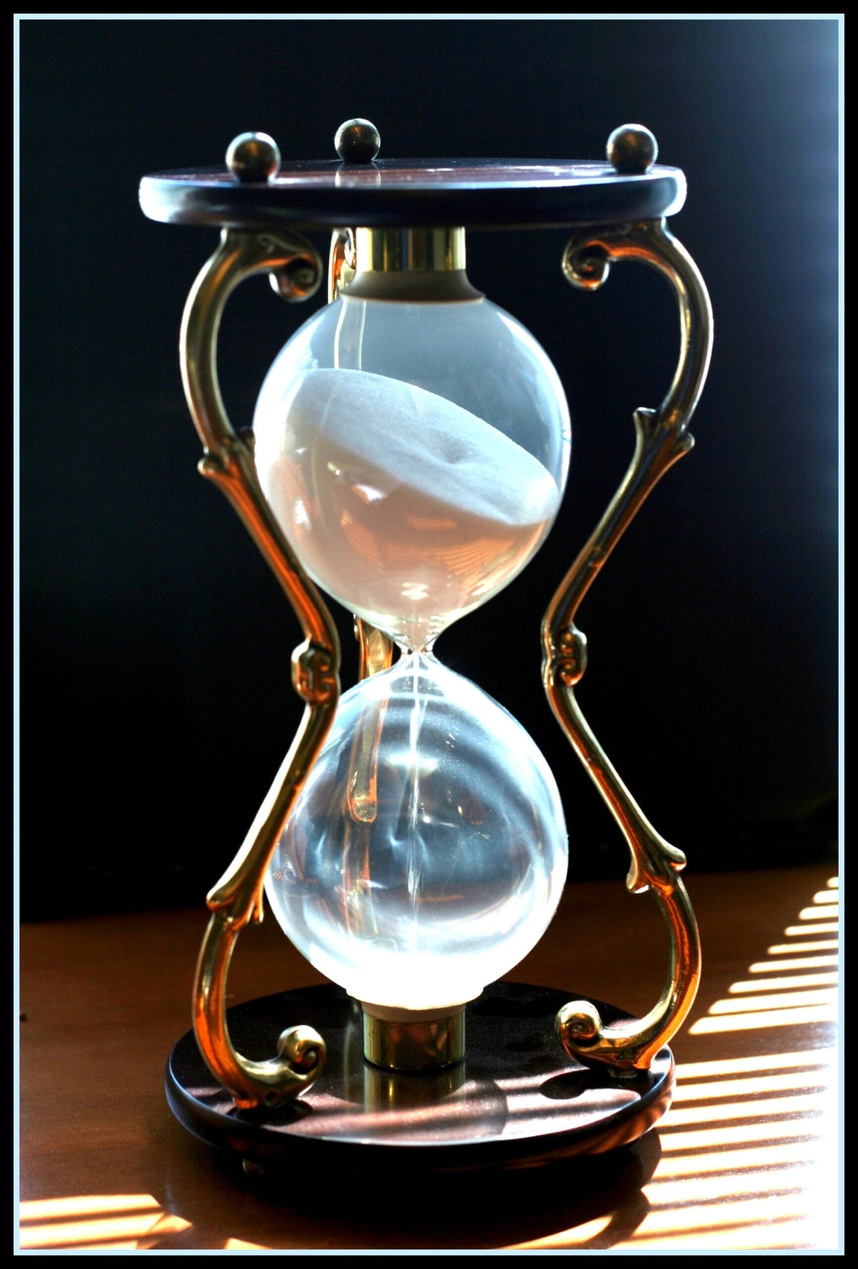 A close-up of a classic hourglass with brass supports, half-filled with white sand, sitting on a wooden surface with sunlight casting striped shadows.