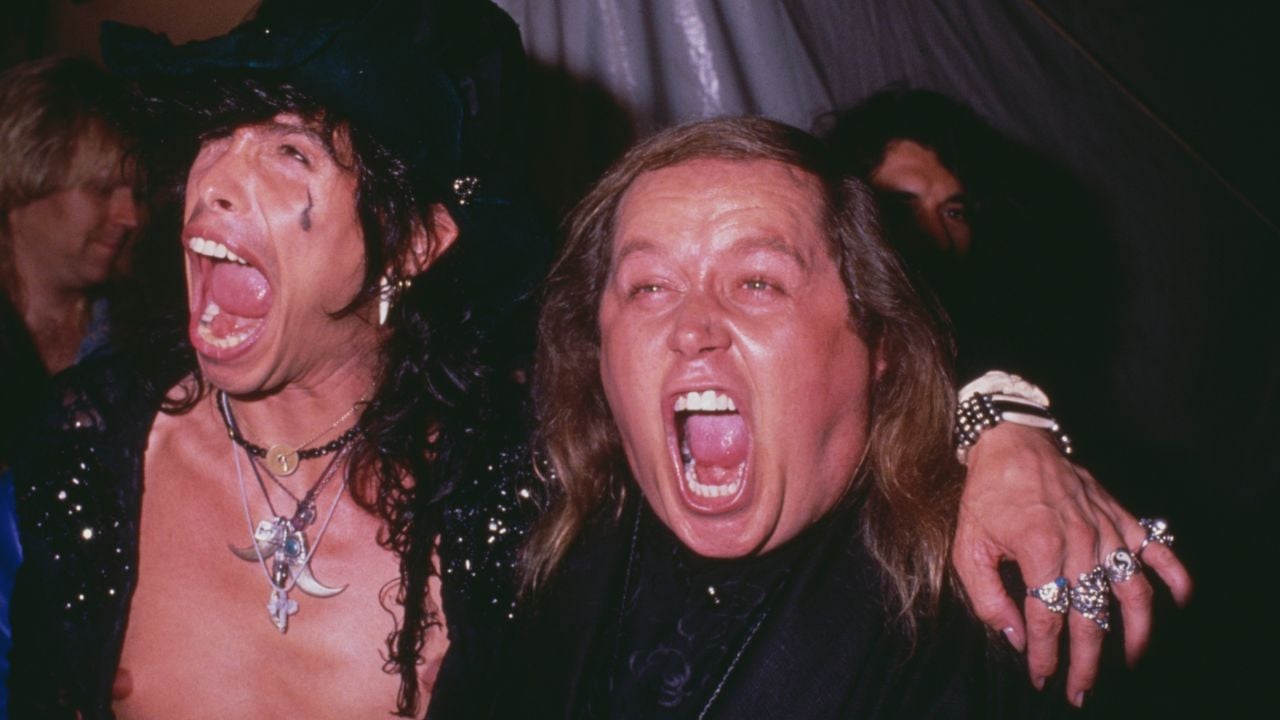 Two people with arms around each other shout or laugh enthusiastically, both showing wide open mouths. One wears a black hat and jewelry, while the other is dressed in dark clothing with rings. They appear to be celebrating together.