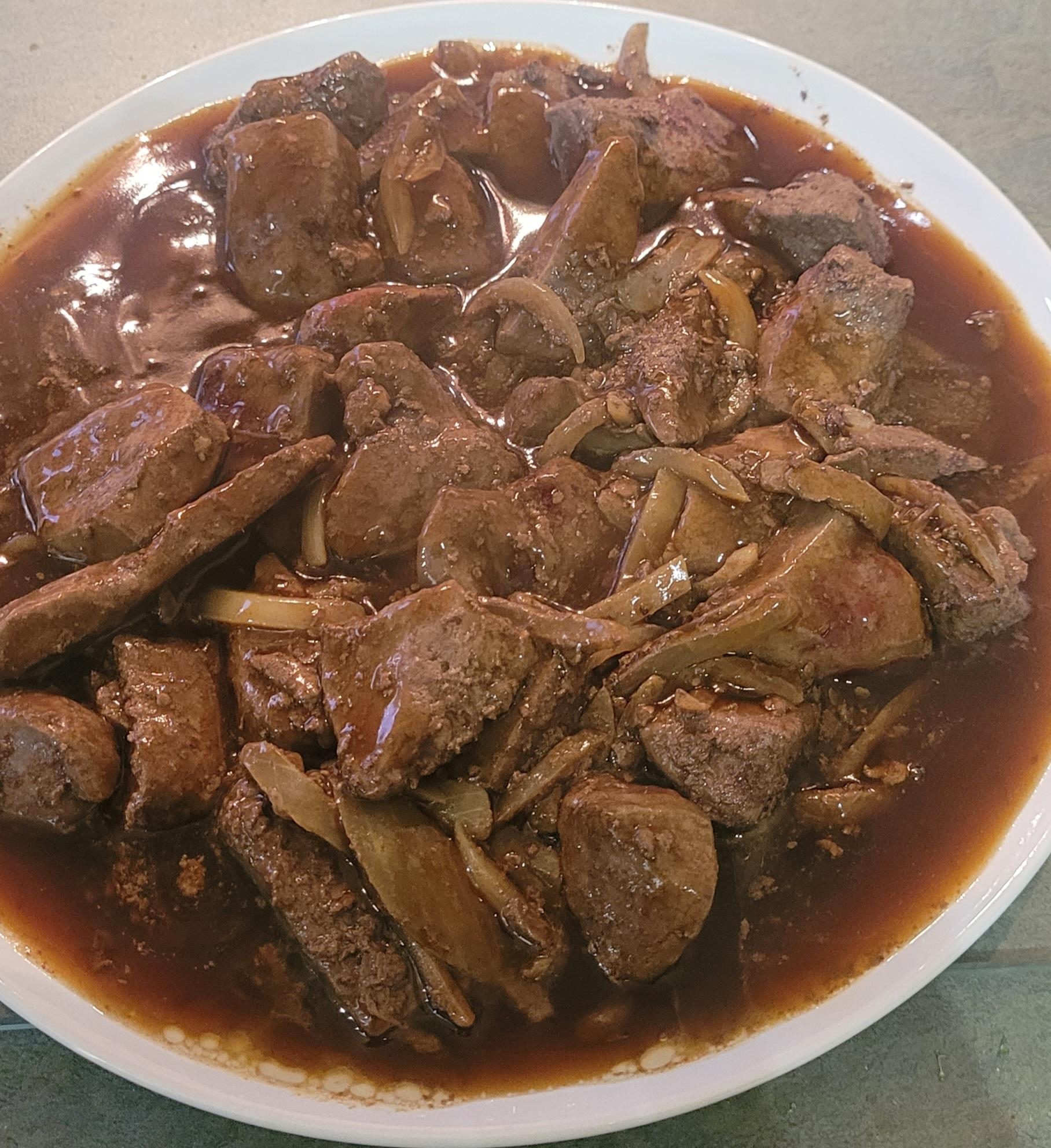 A white bowl filled with chunks of liver and onions in a rich brown sauce, placed on a gray surface.