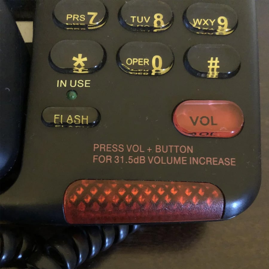 Close-up of a black telephone keypad showing buttons for numbers, flash, and volume. Text reads, “PRESS VOL+ BUTTON FOR 31.5dB VOLUME INCREASE.” A red textured area is visible at the bottom.