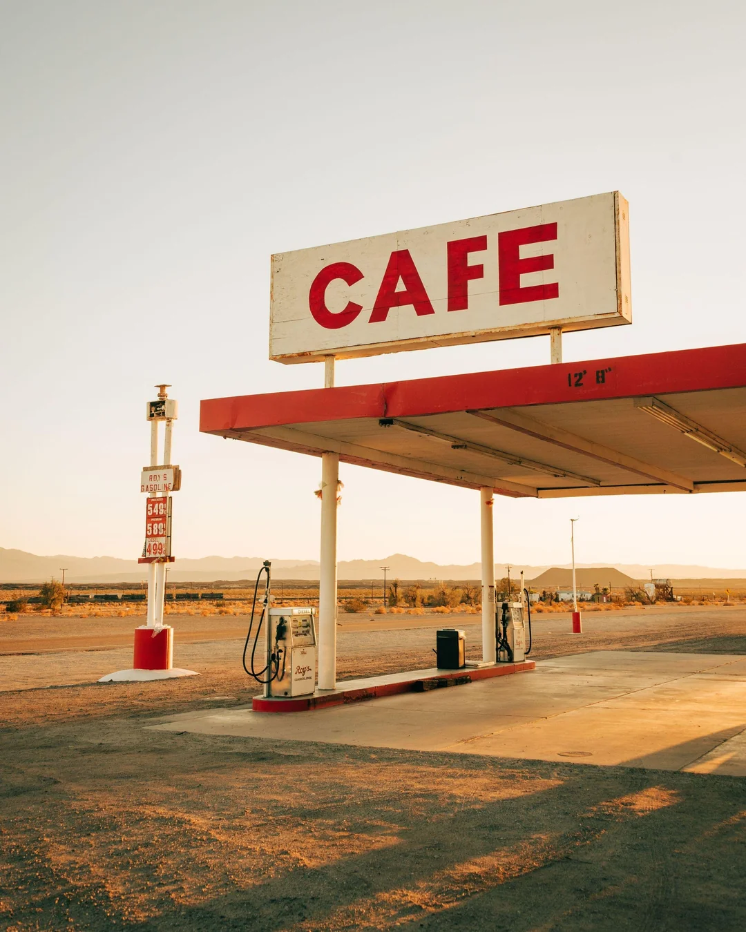 A vintage gas station with an old-fashioned pump and a large red “CAFE” sign, set against a desert landscape at sunset, with mountains in the distance.