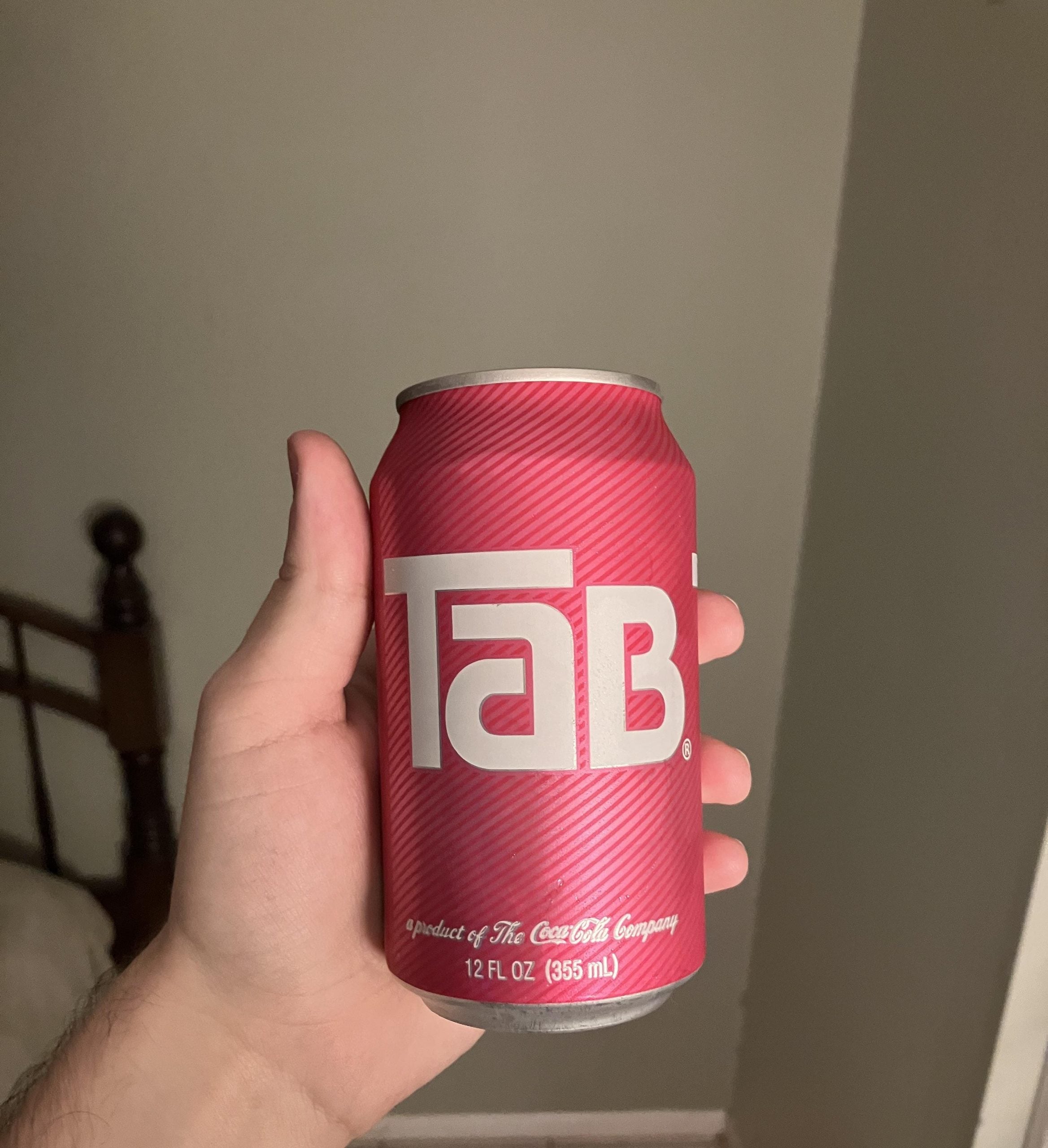 A hand holding a 12 fl oz can of Tab soda with a pink background and bold white logo, next to a wooden bedframe and beige wall in the background.