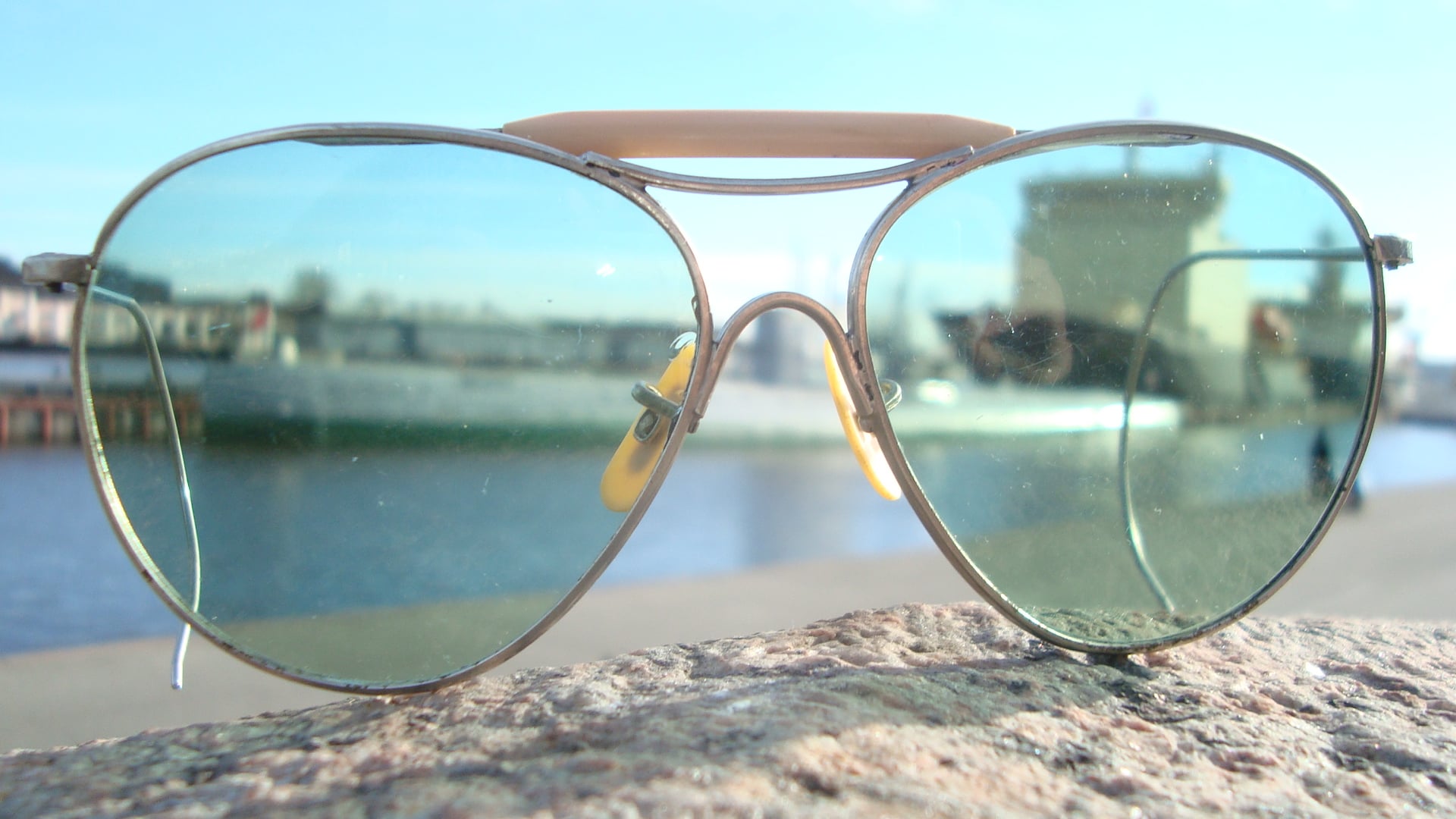 A pair of aviator sunglasses rests on a stone surface, with a blurred view of water and a docked ship in the background, seen through tinted lenses.