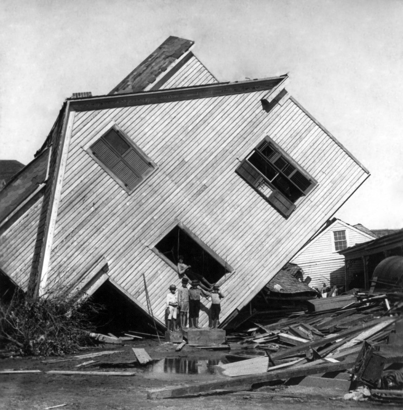 A house tilted dramatically after a disaster, with debris around it. Four children stand in front of the house, looking at the camera. The scene appears chaotic and damaged.