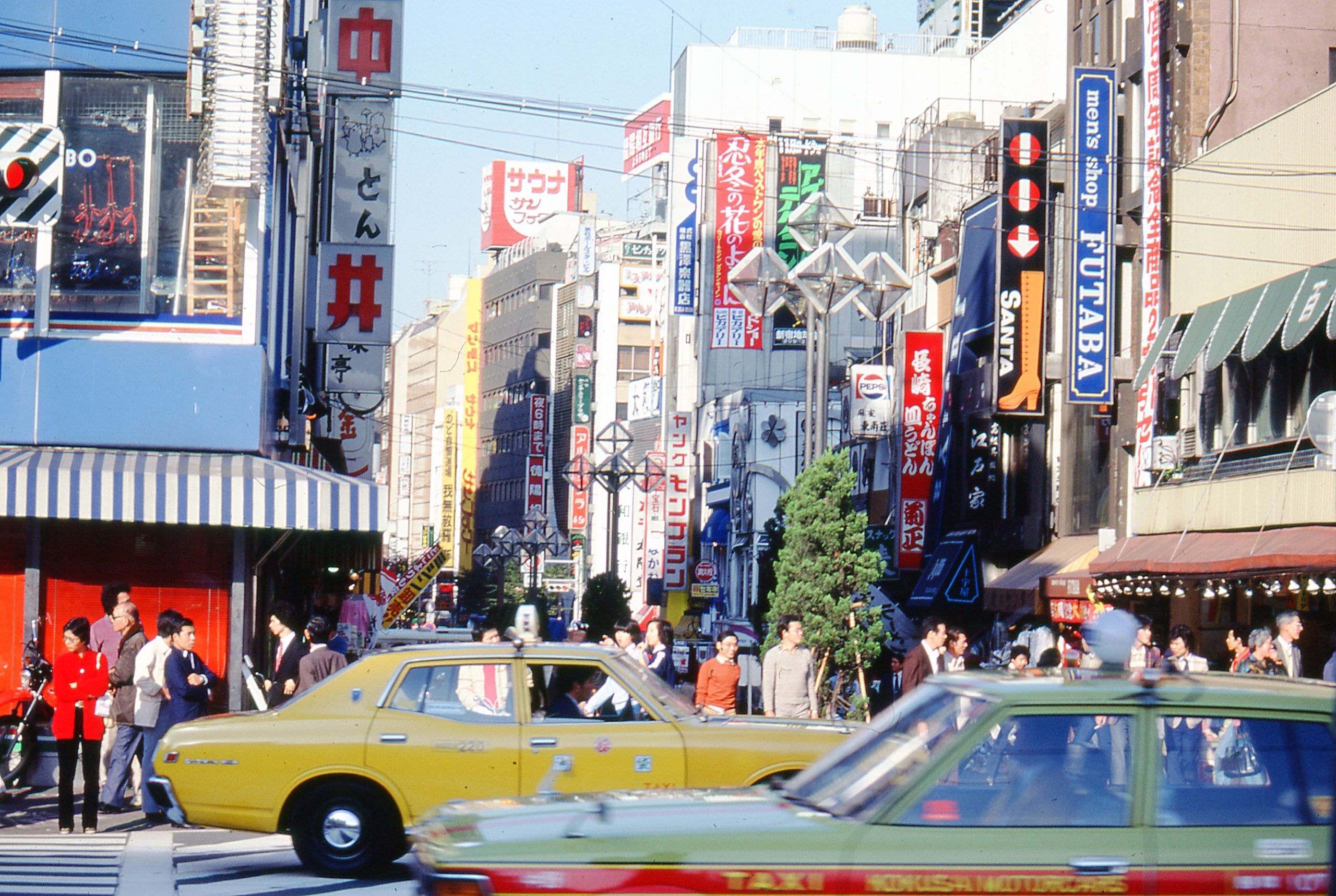 Busy city street in Japan with many people walking, colorful signs in Japanese, and yellow and green taxis driving by tall buildings under bright daylight.
