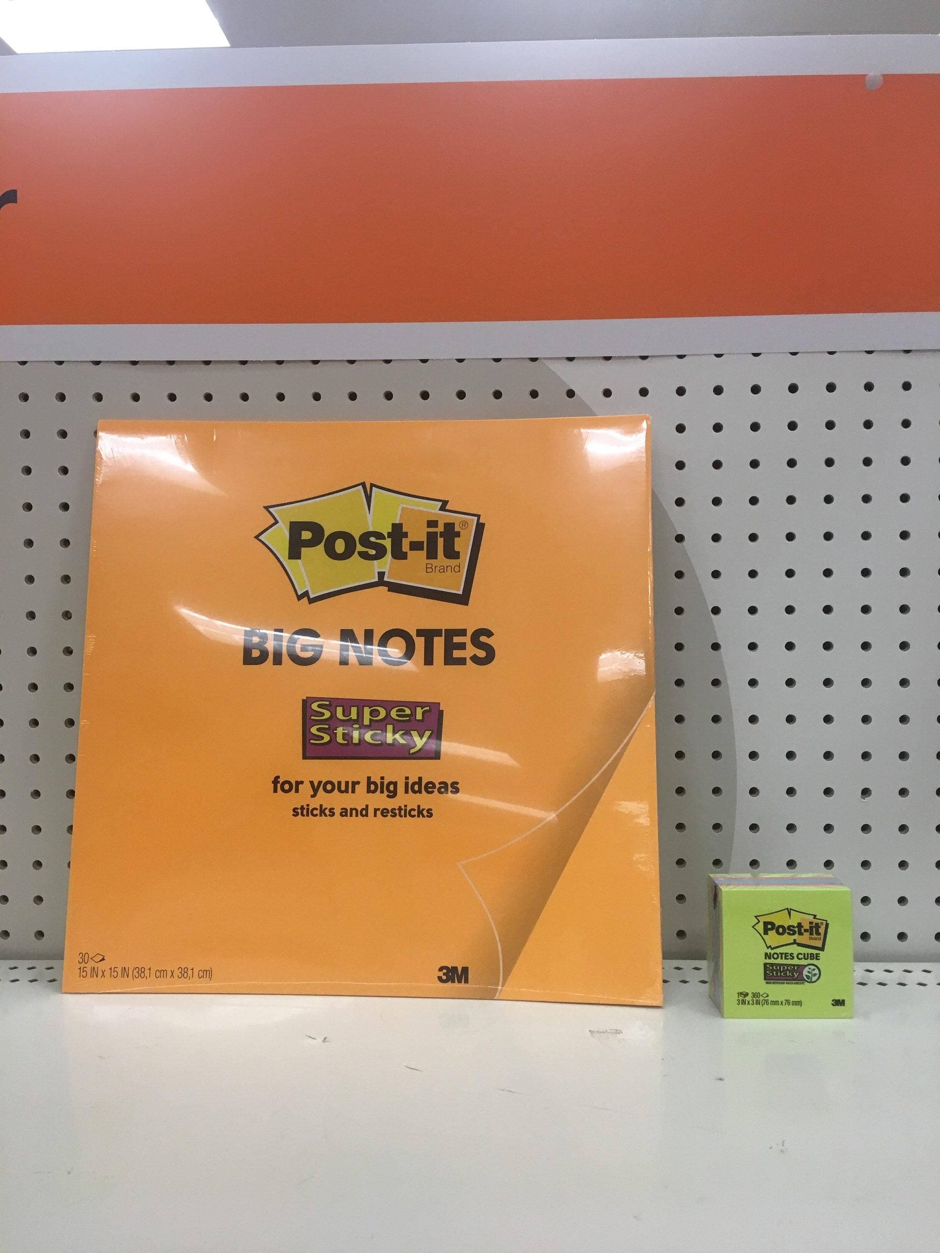Large orange Post-it Big Notes pad is displayed next to a much smaller green Post-it Notes pad on a white shelf with a pegboard background.