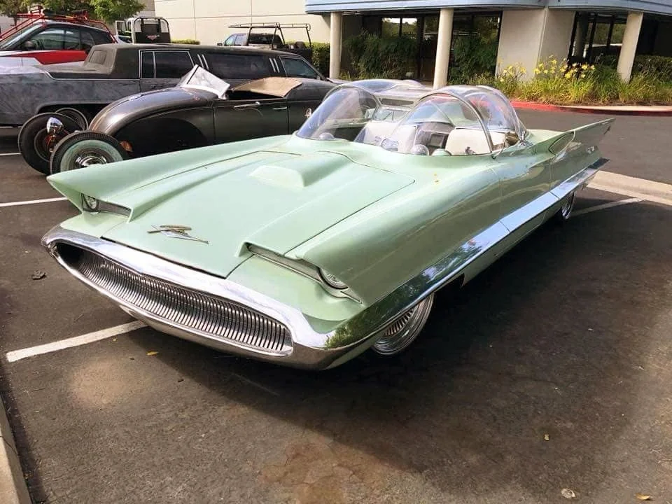 A pastel green vintage concept car with a bubble canopy and chrome details is parked in a lot next to another classic car, outside a modern building.