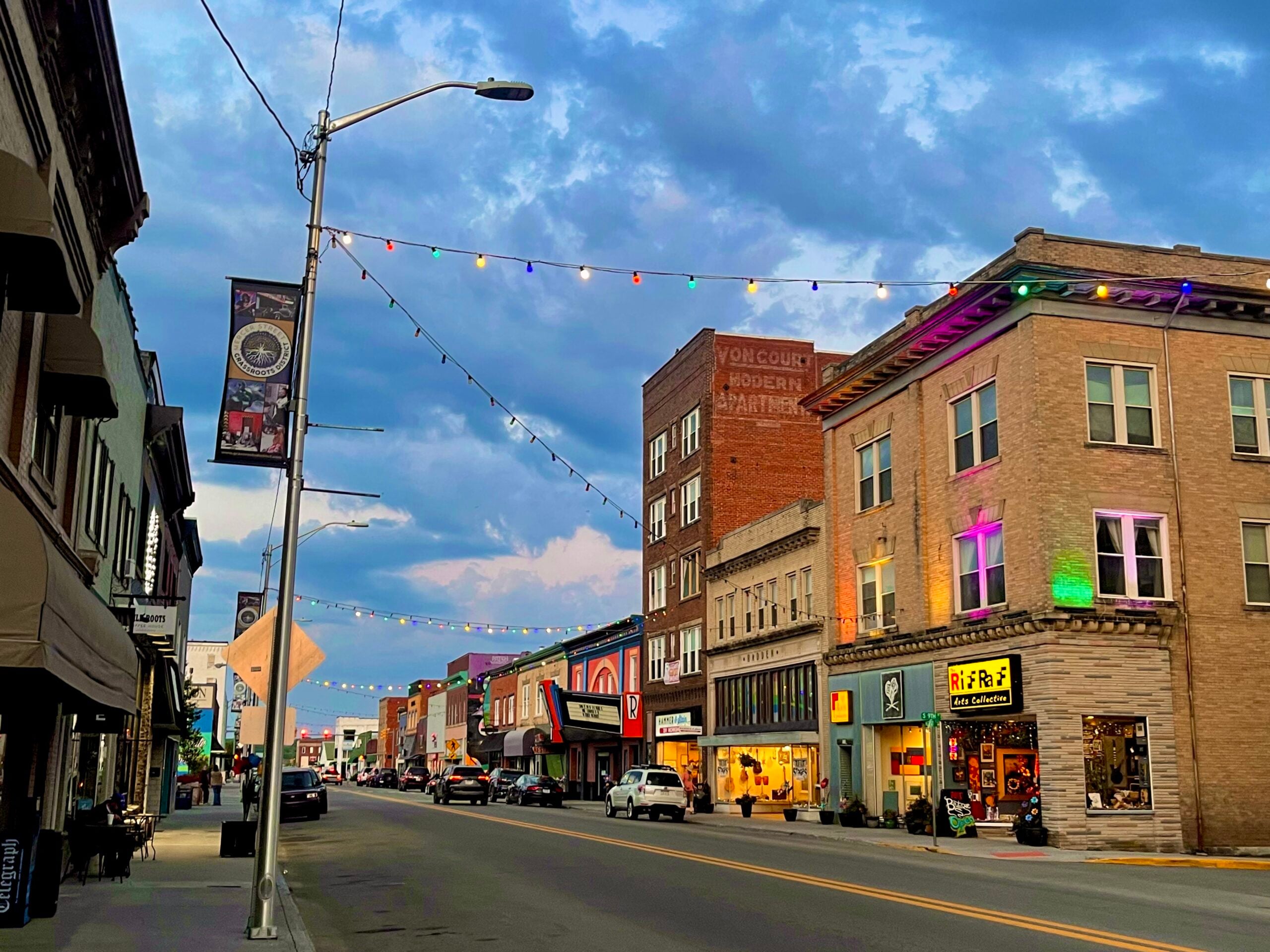 A lively downtown street at dusk with colorful string lights overhead, historic buildings on both sides, shops and restaurants open, and a dramatic cloudy sky above.