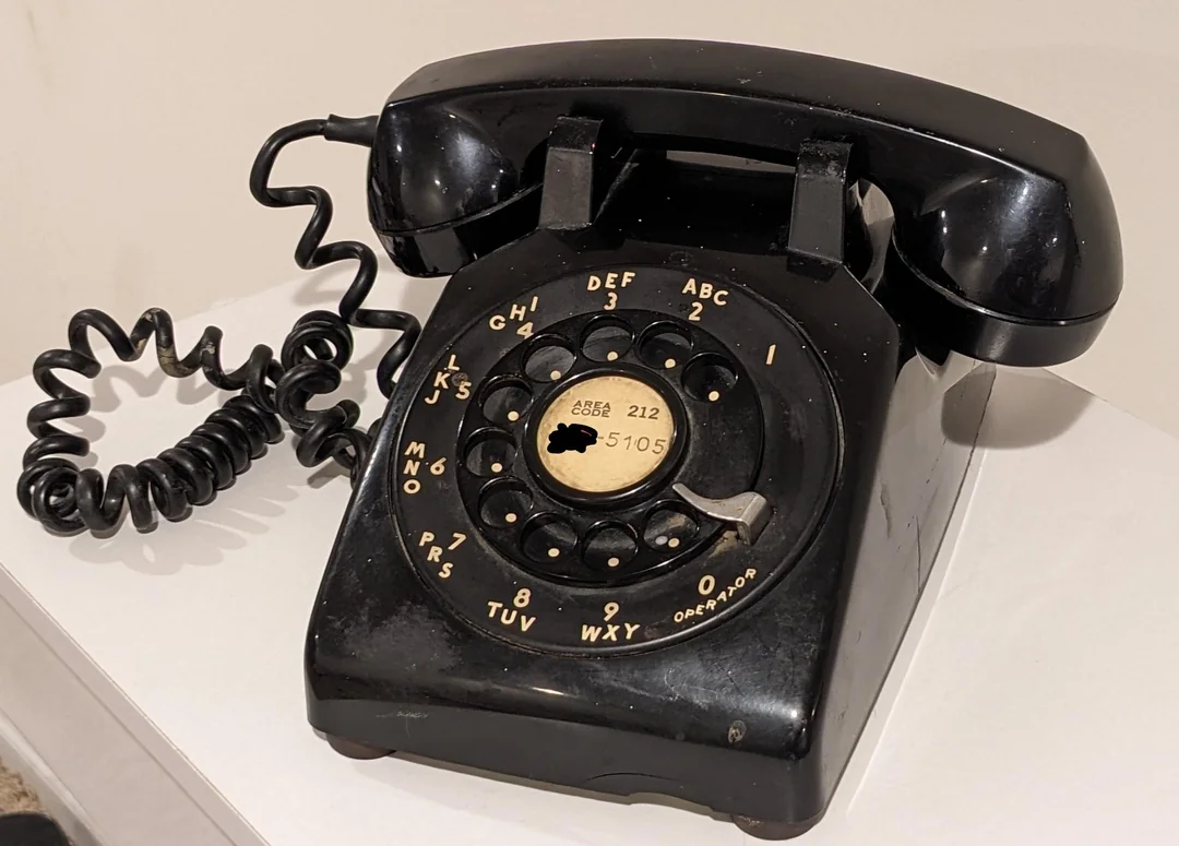 A vintage black rotary dial telephone with a coiled cord sits on a white surface against a plain background.