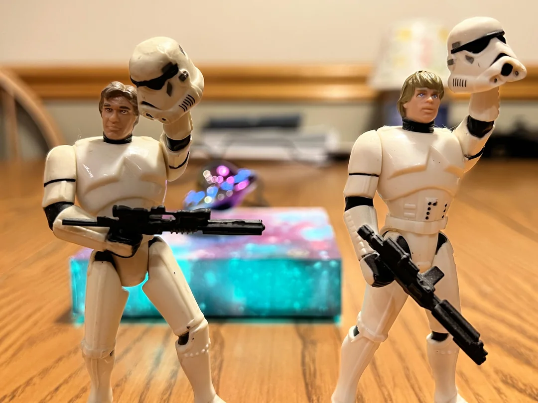 Two Star Wars action figures dressed as stormtroopers stand on a table, each holding a helmet in one hand and a blaster in the other. A colorful box is blurred in the background.
