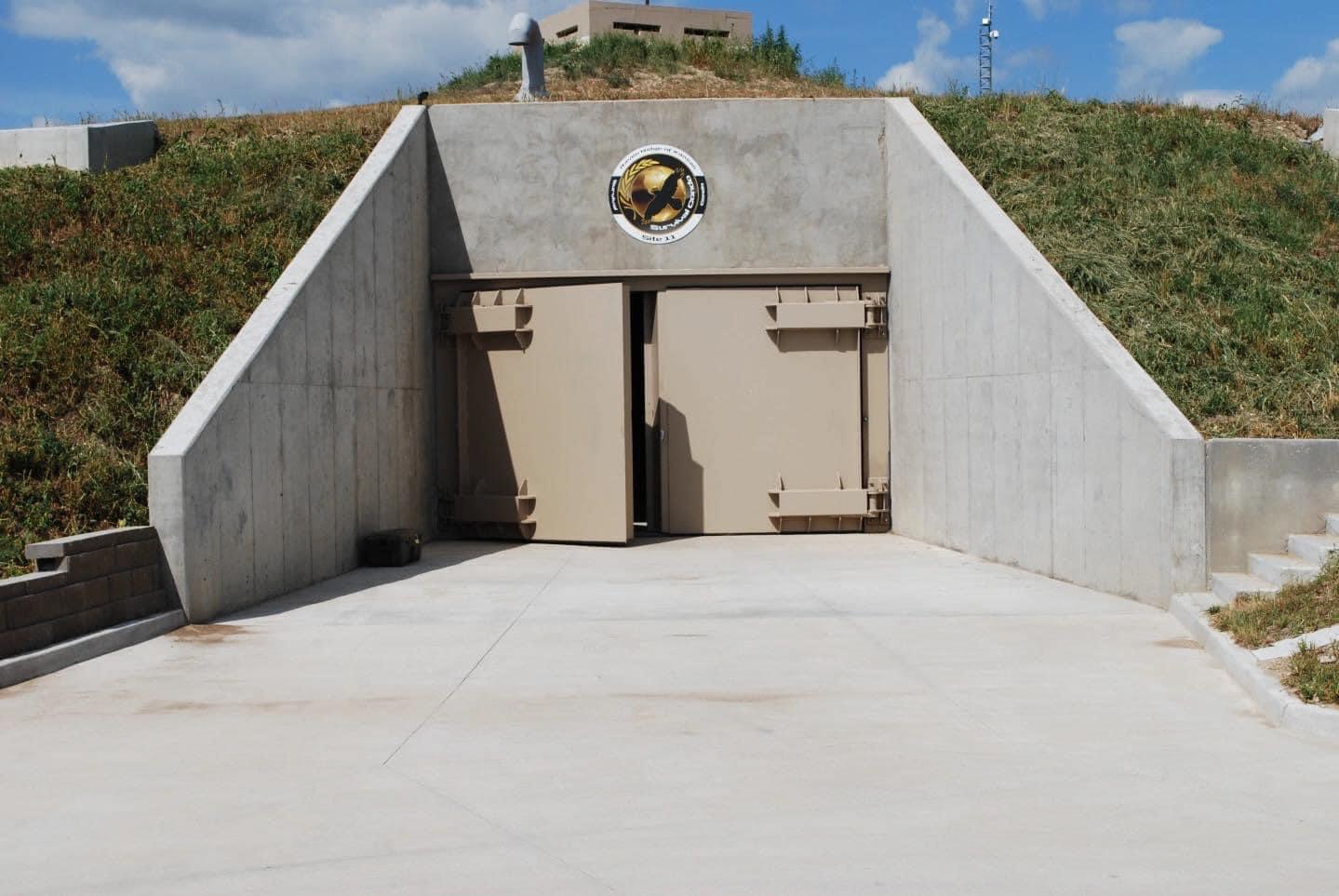 Concrete bunker with large metal doors set into a grassy hillside. A circular emblem is above the entrance. The sky is partly cloudy and the area around the bunker is paved.