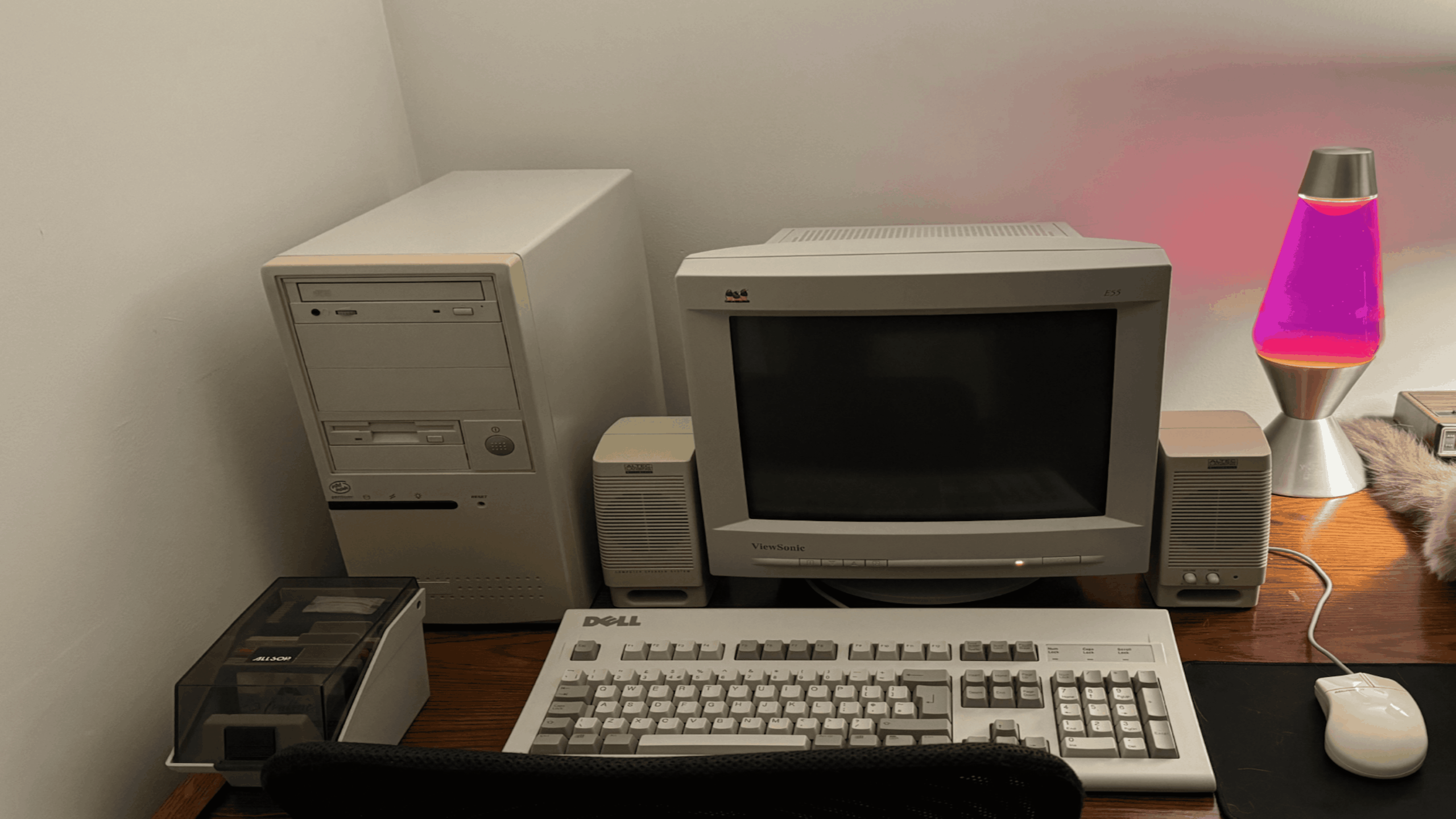 Vintage computer setup with a beige desktop tower, CRT monitor, speakers, floppy disk drive, mechanical keyboard, mouse, and a pink-and-purple lava lamp on a wooden desk against a white wall.