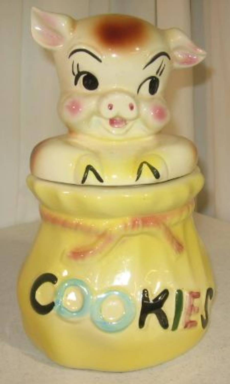 A ceramic cookie jar shaped like a pig’s head on a yellow sack with the word “COOKIES” written in colorful letters.