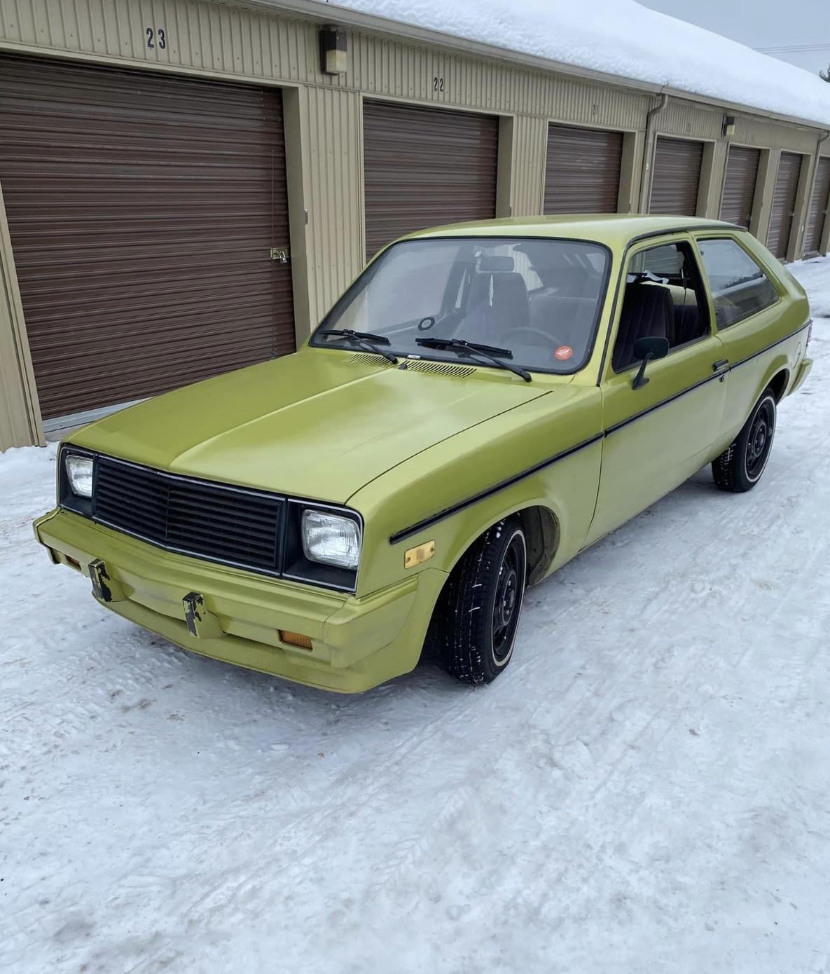 A vintage, light green two-door car is parked on snow in front of tan garage doors. The car appears to be from the late 1970s or early 1980s and shows some minor wear.