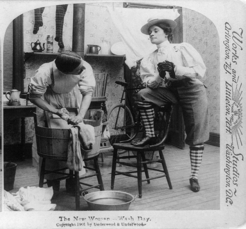 Two women in early 1900s attire are in a kitchen; one washes clothes by hand, while the other confidently poses with a pipe, foot on a chair, wearing striped socks, bloomers, and a hat.